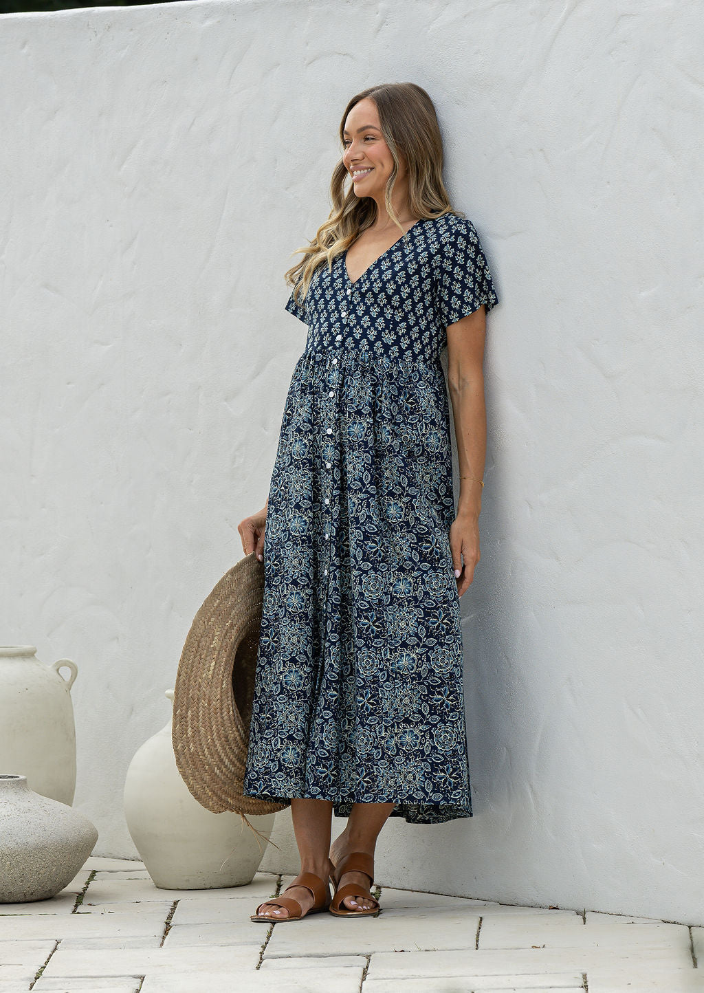Woman wearing a blue floral dress standing against a white wall with decorative vases.