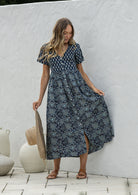 Woman standing in a 100% cotton floral print dress with a V-neckline, short sleeves, pockets, and buttons through to the knee.
