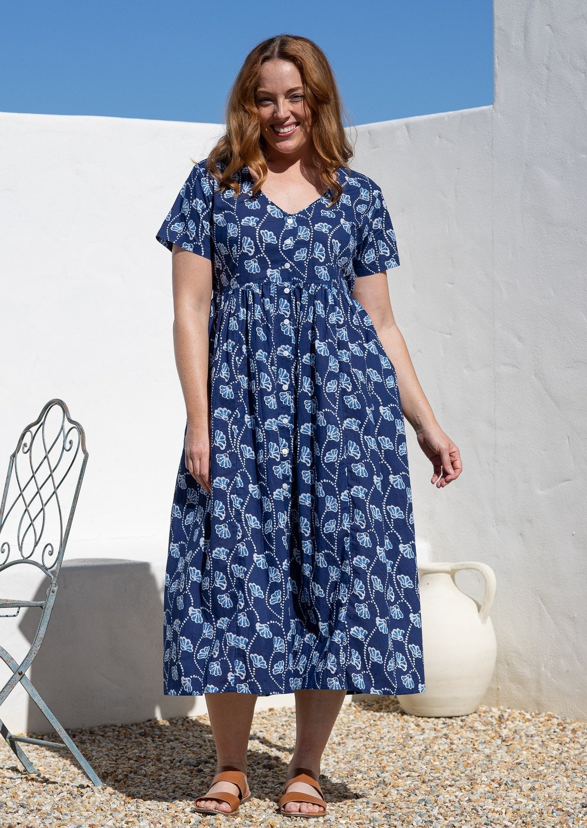 A woman wearing a navy blue cotton midi dress with a white botanical print. The dress has short sleeves, a V-neckline, and is button-through. 