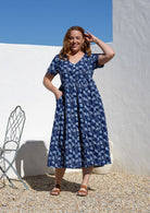 Woman standing in a 100% cotton floral print dress with a V-neckline, short sleeves, pockets, and buttons through to the knee.