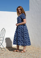 Woman stands sideways wearing a 100% cotton dress with short sleeves, pockets, and a drawstring tie at the back.