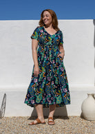 A woman wearing a short-sleeve V-neck cotton midi dress with a colourful floral and peacock print on a dark navy background, with hand in pocket