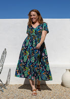 A woman wearing a short-sleeve V-neck button through cotton midi length dress with a colourful floral and peacock print on a dark navy background, with a shoestring ties cinching the waist