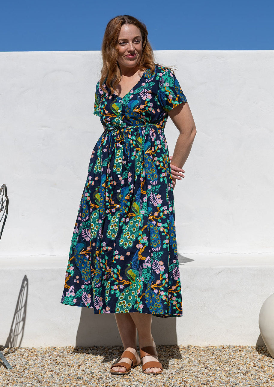A woman wearing a short-sleeve cotton midi dress with a colourful floral and peacock print on a dark navy background, standing outdoors against a white wall.