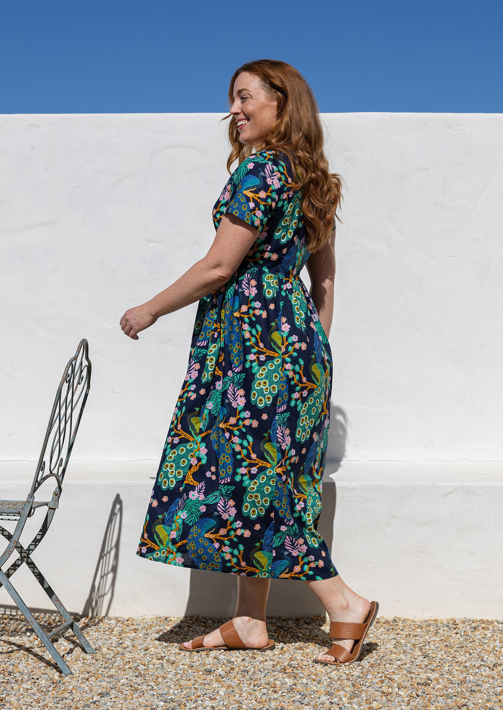 Woman standing side on wearing a short-sleeve cotton midi length dress with a floral and peacock print on a dark navy background, walking outdoors against a white wall under a blue sky.