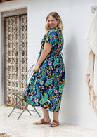 A woman stands sideways wearing a short-sleeve V-neck button through cotton midi length dress with a shoestring ties cinching the waist