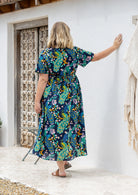 Woman standing backwards wearing a short-sleeve cotton midi length dress with a floral and peacock print on a dark navy background. 