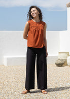 Model stands in plain sleeveless cotton top and black pants against a white wall and a blue sky