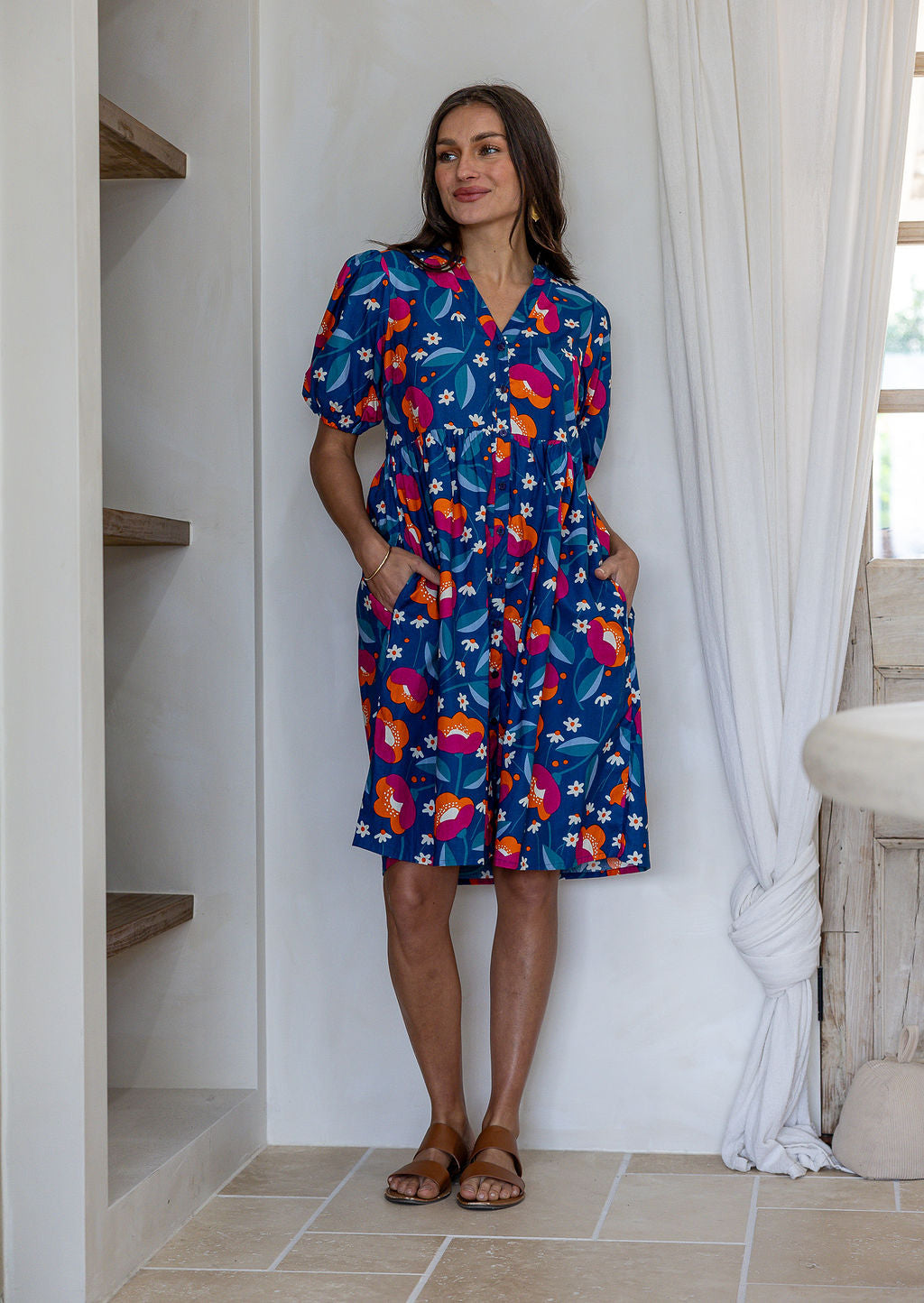 Woman wearing a knee length relaxed fit button through 100% cotton dress with elbow length sleeves and hand in pockets.