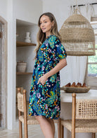 Woman with one hand in pocket standing sideways wearing a 100% cotton dress with white multi colour floral print on a navy base