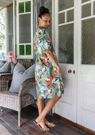 Woman stands sideways wearing a colourful dress standing on a porch with wicker furniture and a white door.