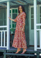 Woman stands side ways wearing a 100% cotton tiered maxi dress in cream based floral print with 3/4 sleeves, pockets and mandarin collar