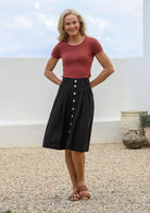 Woman posing in knee length black cotton button-up skirt with belt loops, pockets and box pleats