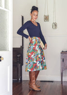 Woman wearing a blue top and colorful skirt in a room with wooden floors and white walls.