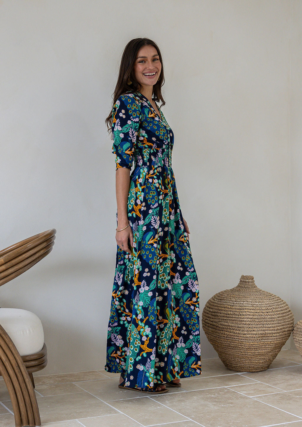 A woman standing side on wearing a navy blue rayon maxi dress with 3/4 sleeves, buttoned bodice and a wide shirred waistband, in a colourful floral and peacock print