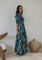 A woman standing side on wearing a navy blue rayon maxi dress with 3/4 sleeves, buttoned bodice and a wide shirred waistband, in a colourful floral and peacock print