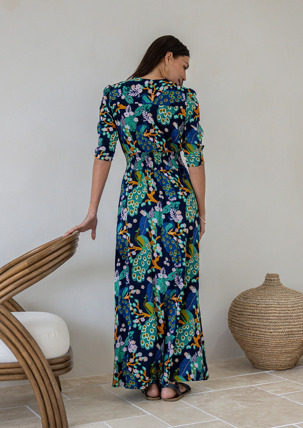 Back view of woman wearing a 3/4 sleeved rayon maxi dress with a vibrant floral and peacock print in shades of teal, green, blue, orange, and purple on a dark navy base.