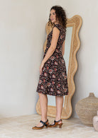 Woman stands sideways wearing a floral cotton dress with cap sleeves, piped details 