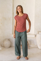 Woman wearing a 100% rayon terracotta shell t-shirt and green pants standing in a room with white walls and decorative vases.