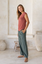 Woman stands sideways wearing a 100% rayon terracotta shell t-shirt and green pants standing in a room with white walls and light-colored floor.