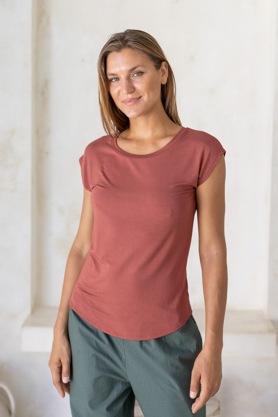 Woman wearing a 100% rayon terracotta shell t-shirt and green cotton pants against a white wall.