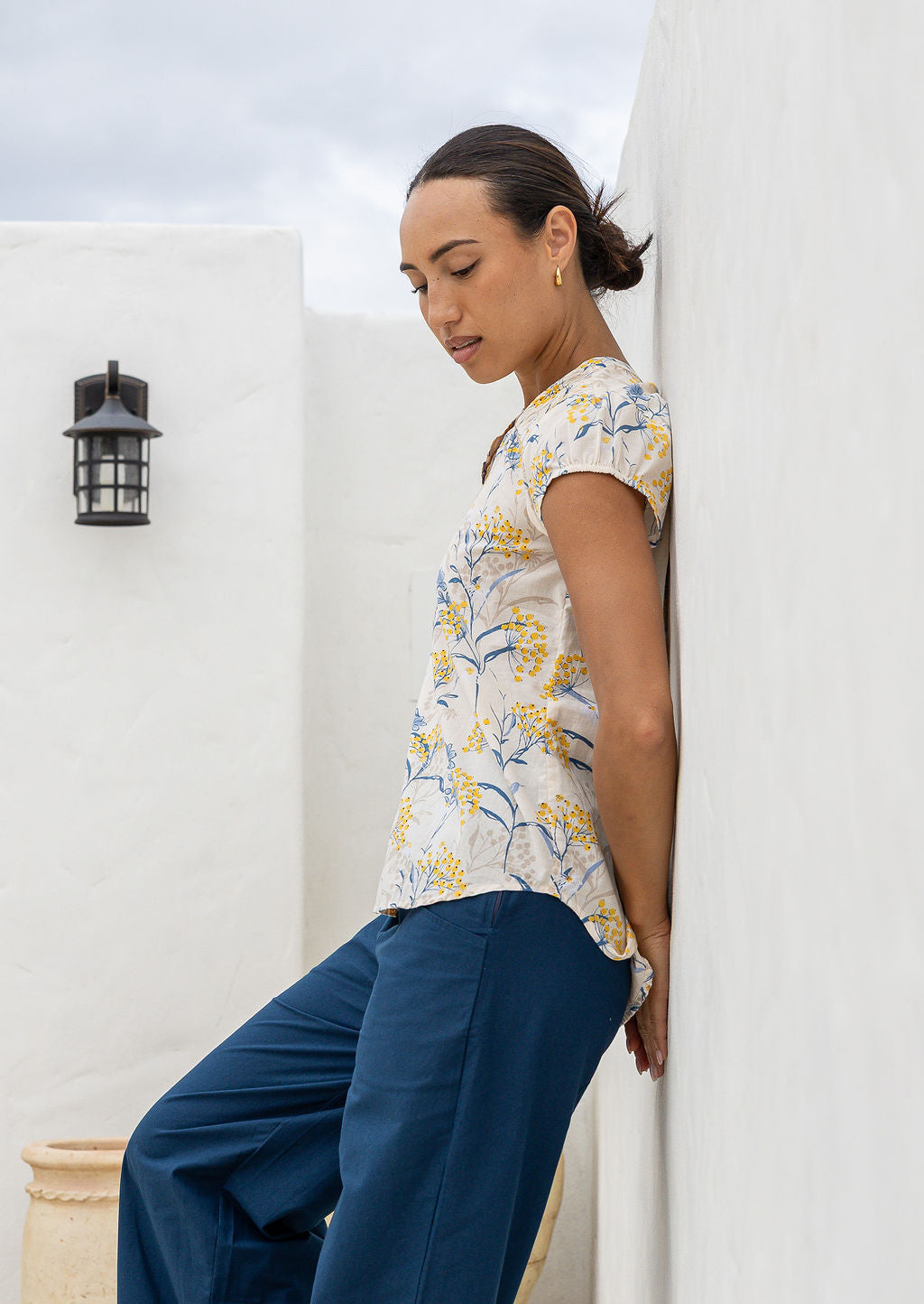 Woman wearing a 100% cotton neutral based floral blouse and blue pants leaning against a white wall.