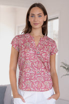 Woman wearing a warm pink based floral  blouse and white pants standing against a white wall.