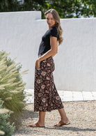 Woman stands sideways both hand in pockets wearing a black based floral print wide leg, 3/4 length cotton pants with side zipper 