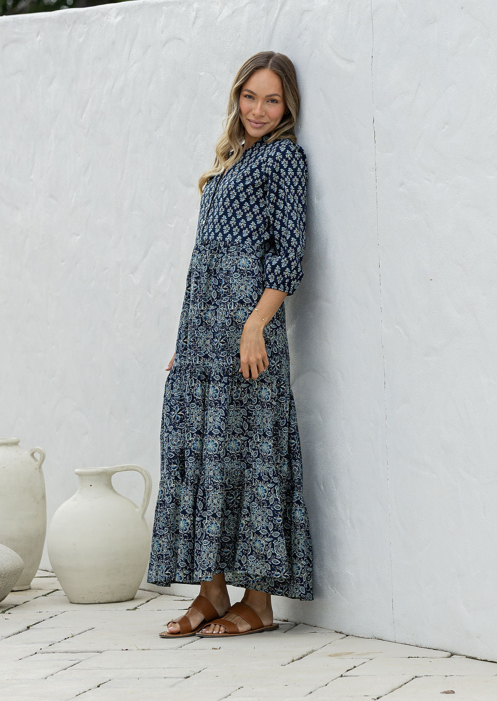 Woman wearing a 100% cotton tiered maxi dress in navy blue based floral print against a white wall with white vases in the foreground.