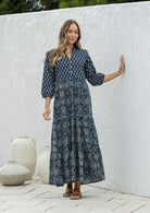 Woman standing in navy blue based 100% cotton flowy tiered maxi dress with 3/4 sleeves, buttoned bodice and pockets 