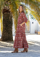 Woman stands side ways wearing a 100% cotton tiered maxi dress in red based floral print with 3/4 sleeves, pockets and mandarin collar
