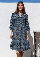 Woman wearing a blue patterned dress and brown boots standing against a white wall.