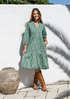 Woman wearing a sage green based floral dress and brown boots standing in front of a white wall.