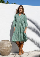 Woman in a floral dress with pockets and brown boots standing in front of a white wall.