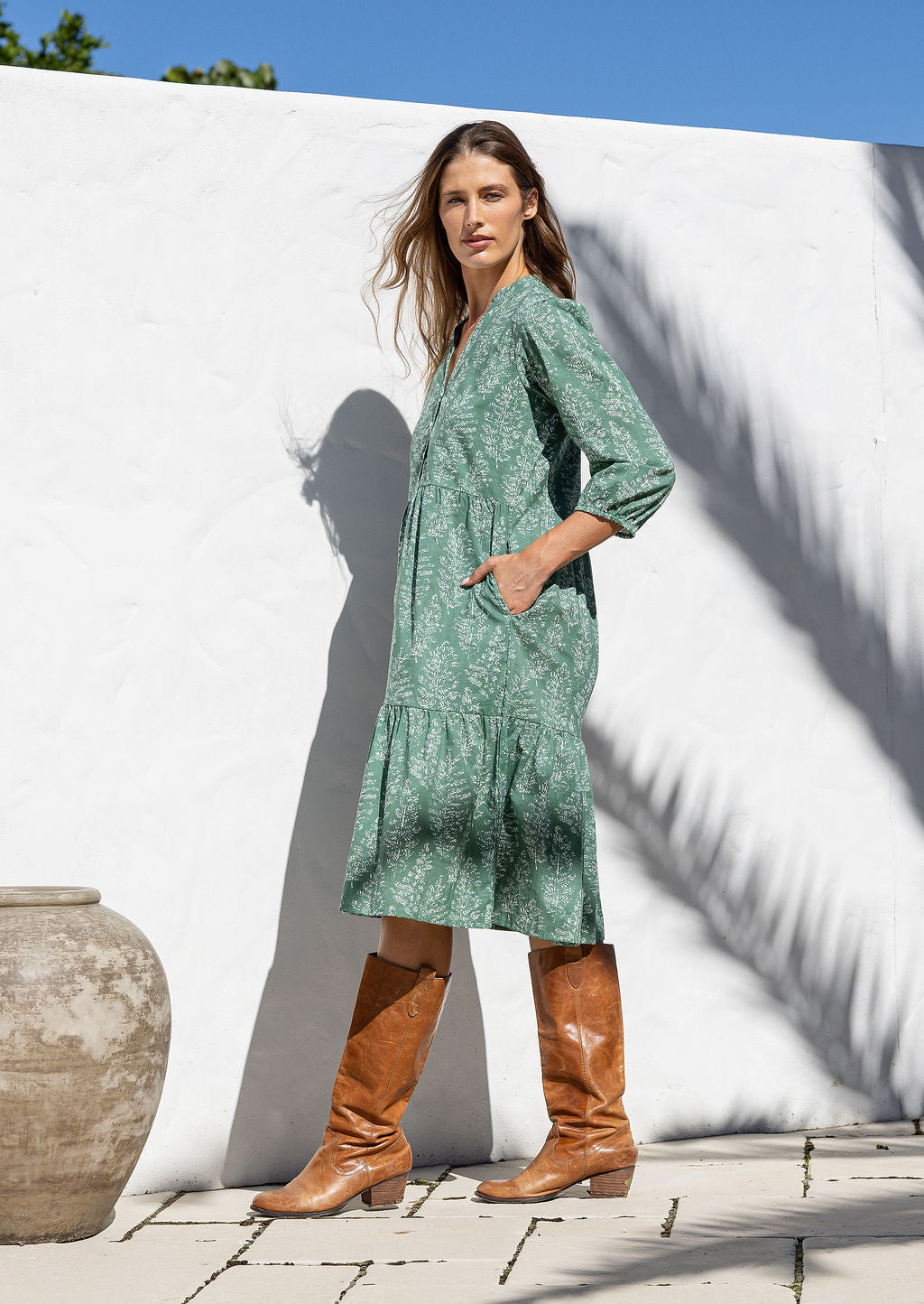 Model stands side on in cotton over the knee length relaxed fit dress with 3/4 sleeves, paired with brown boots. 