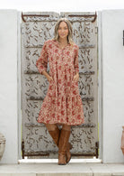 Woman wearing a red floral dress and brown boots standing in front of a decorative wooden door.