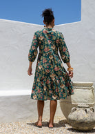 Model stands backwards in cotton tiered over the knee floral print dress, holding the skirt out, paired with tan sandals