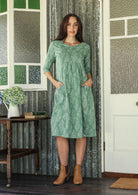 Woman wearing a 100% cotton sage green based floral dress with sleeves and hand in pockets