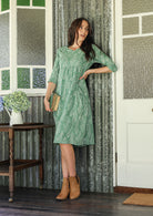 Woman wearing a cotton sage green based floral dress with sleeves standing in a rustic room