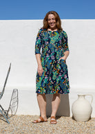 A woman wearing a knee-length, three-quarter sleeve cotton dress with a vibrant multicolour floral and peacock print on a dark navy background, standing outdoors against a white wall.