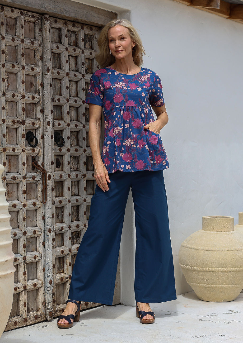 Woman standing with hand in top's pocket, wearing a lightweight 100% cotton blue floral print top with dark blue cotton pants