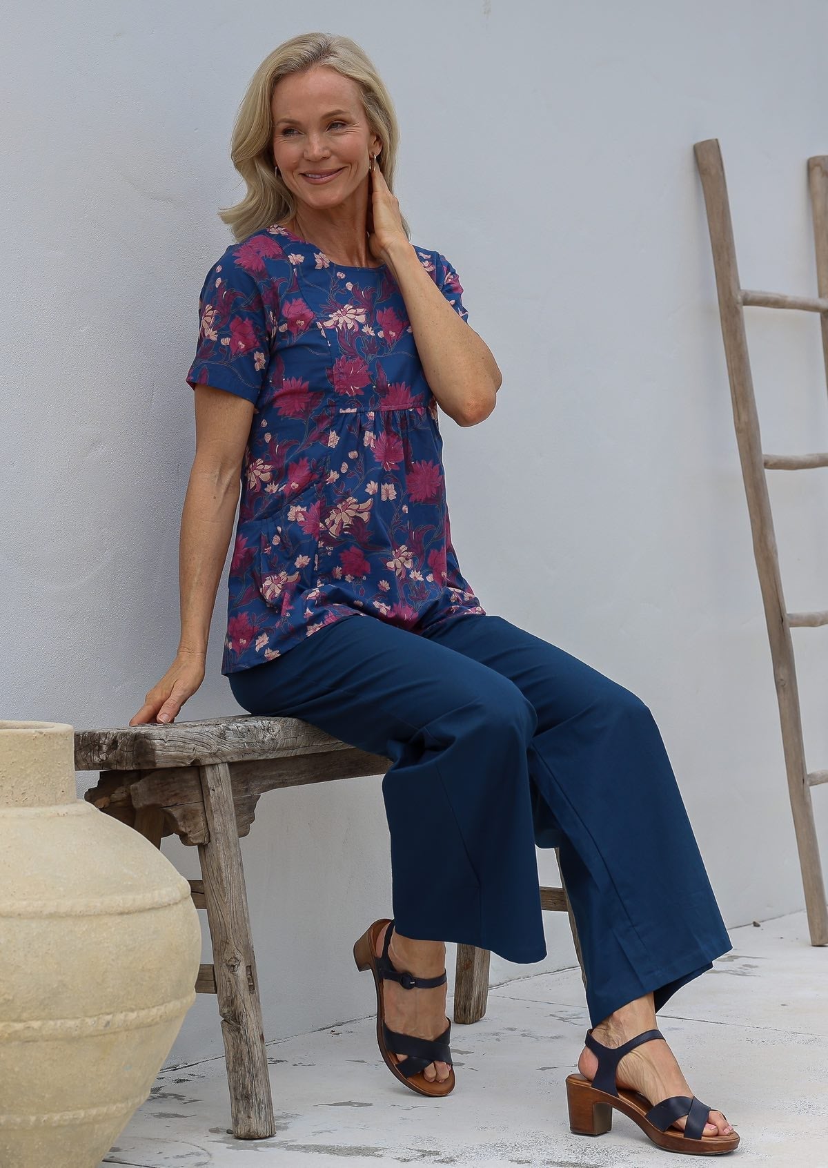 Woman sitting on a wooden bench wearing a blue floral cotton top with short sleeves, round neckline, empire waist and pockets