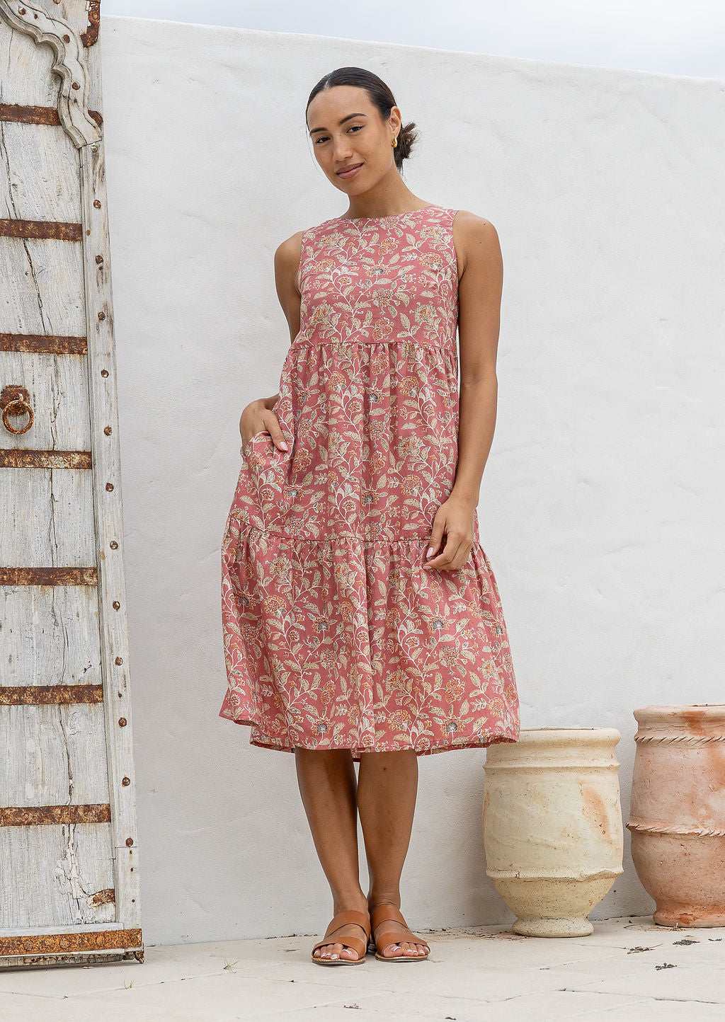 Woman in a sleeveless, floral rose pink based 100% cotton dress standing in front of white wall. 