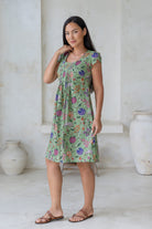 Woman wearing a green floral A-line dress with fitted bodice, cap sleeves and round neckline, standing in a room with white walls and a vase.