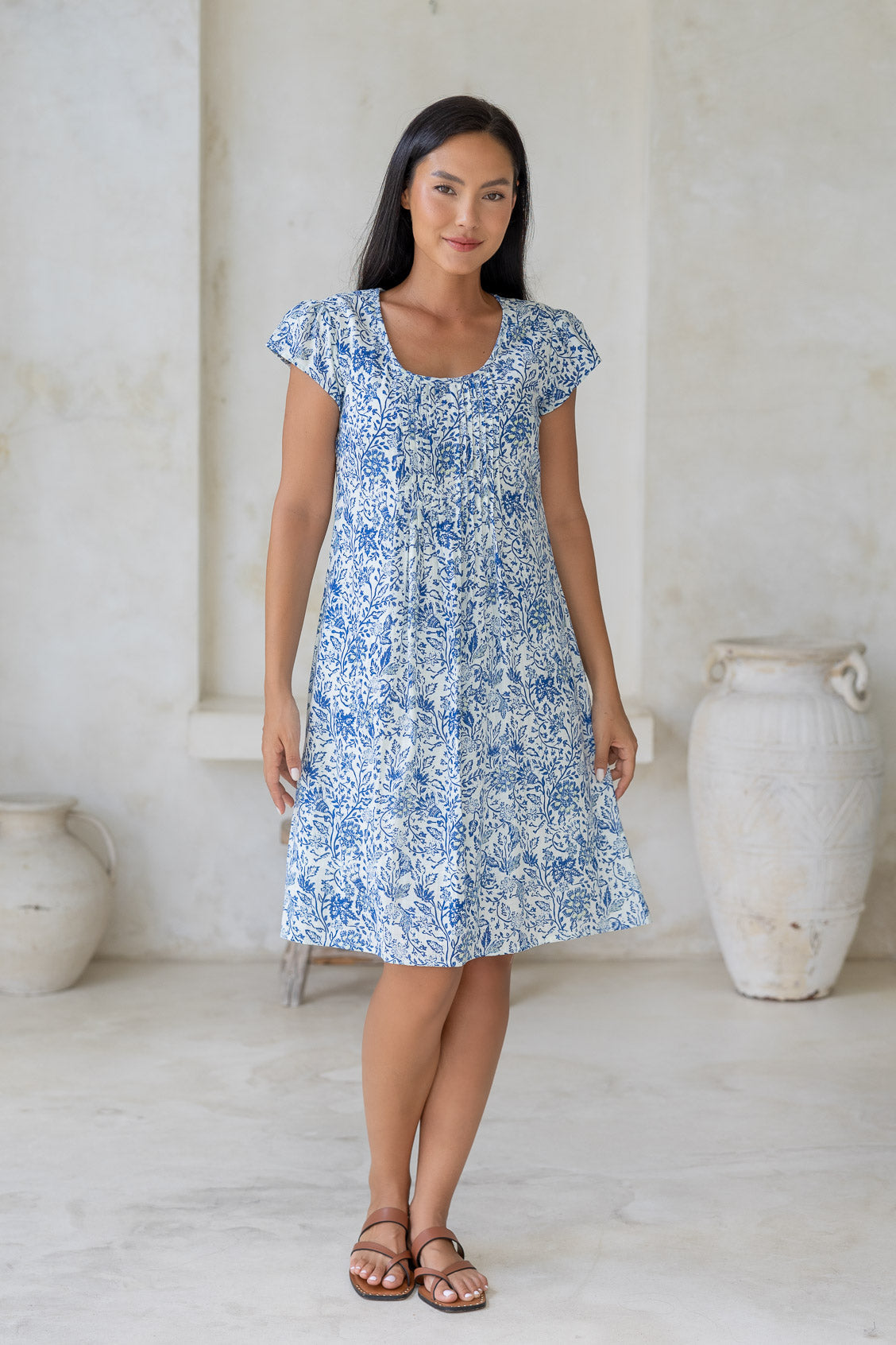 Woman wearing a cap sleeved, above knee length, white based cotton dress with blue floral print and lemon yellow highlights.