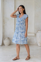 Woman wearing lightweight cotton dress with round neckline, small pleats across the bodice and cap sleeves in white based blue floral print.