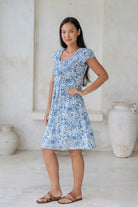 Woman posing in 100% cotton above knee length dress with round neckline, cap sleeves and small pleats across the bodice in white based floral print.