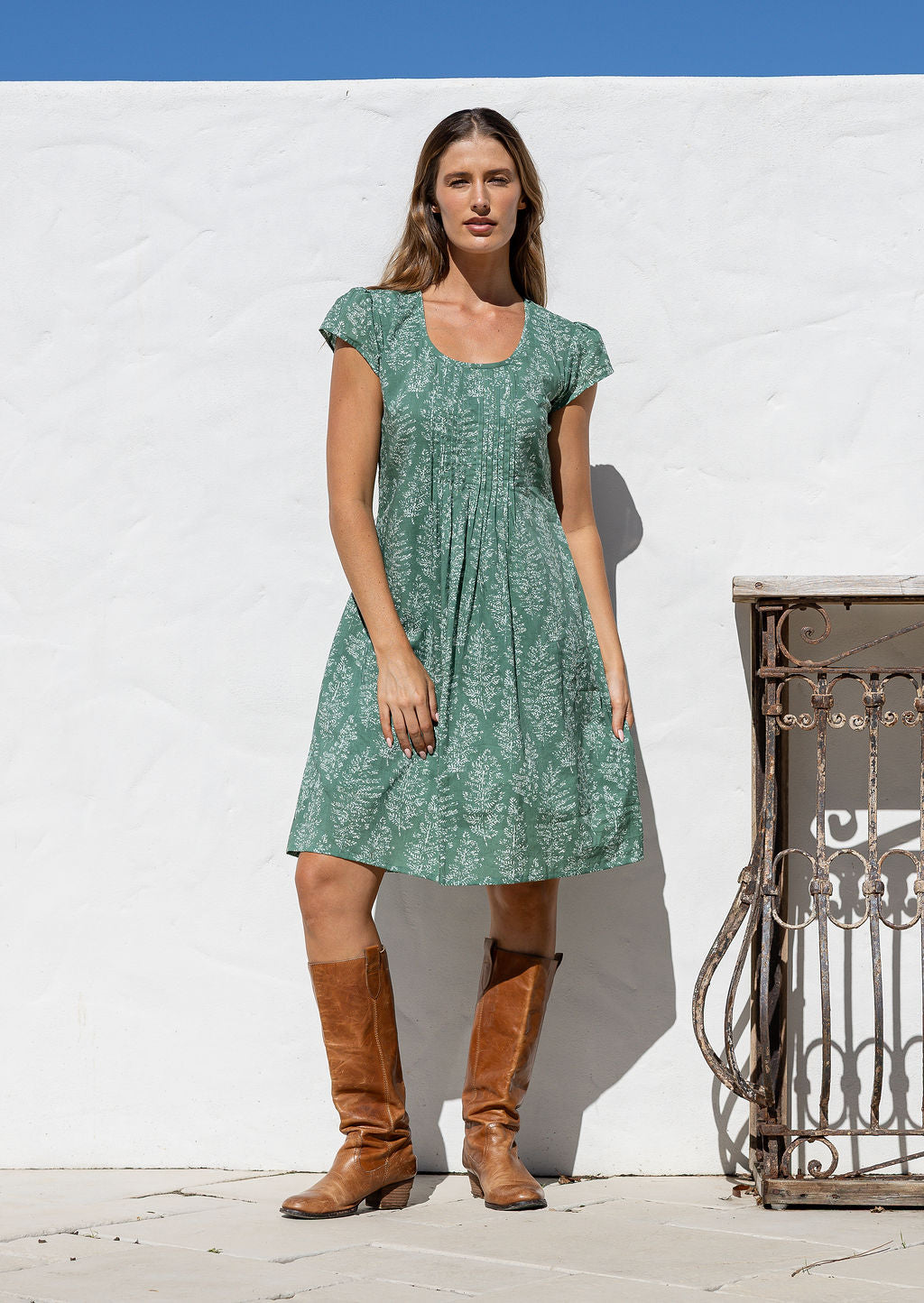 Woman wearing a 100% cotton sage green based floral dress standing in front of a white wall. 