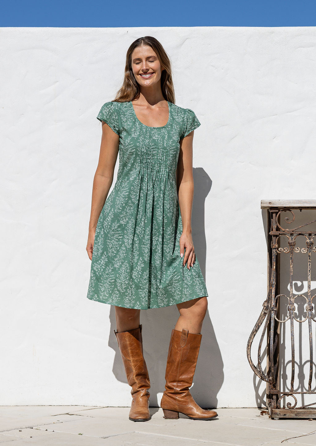 Woman wearing a green floral dress and brown boots standing against a white wall.