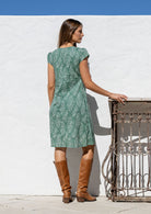 Woman stands backwards wearing a 100% cotton sage green based floral dress with round neckline and cap sleeves.